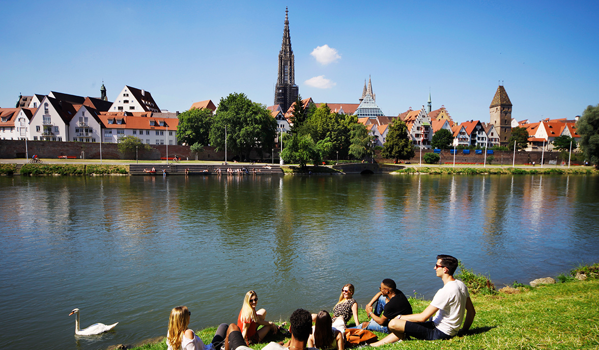 Bild zeigt junge Menschen die an der Donau sitzen. Im Hintergrund sieht man das Ulmer Münster 