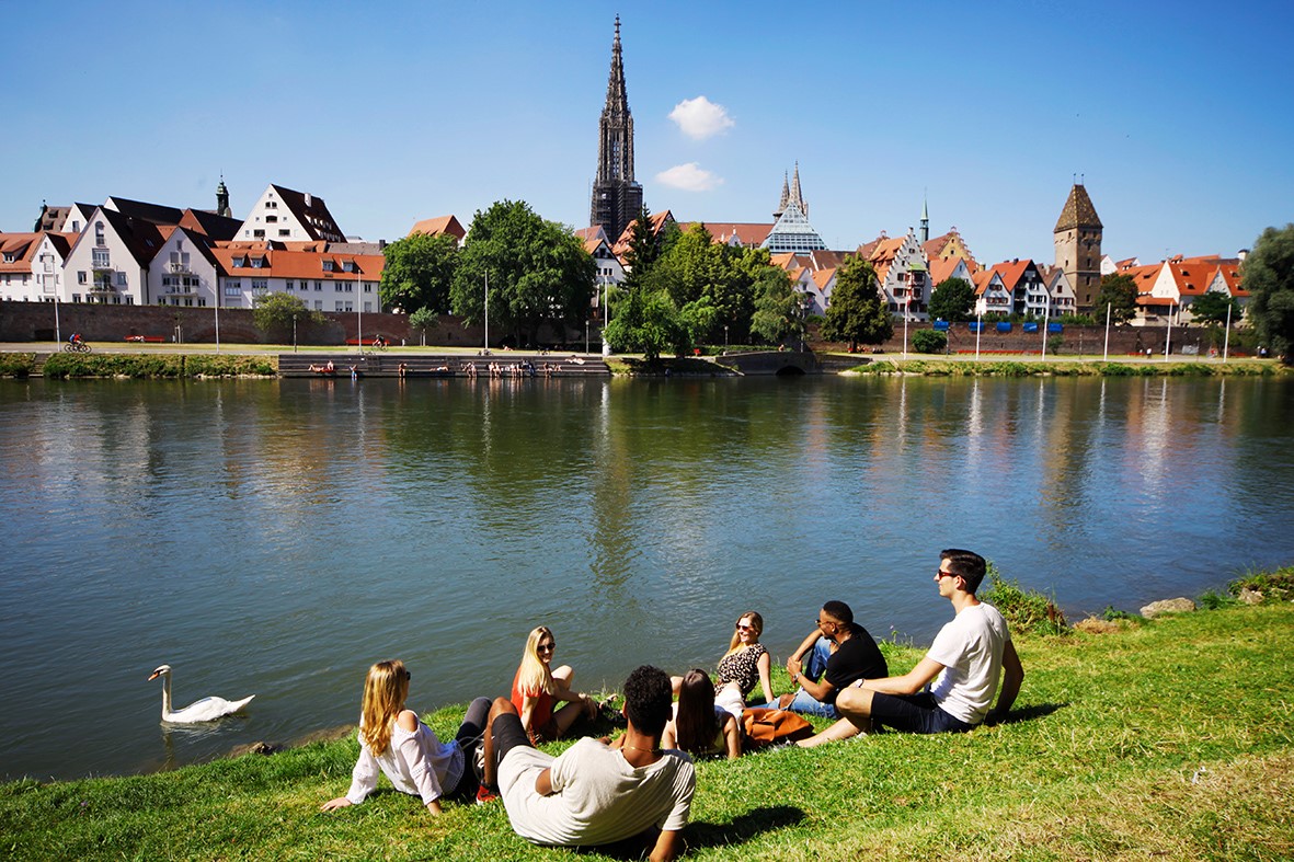 Junge Menschen die an der Donau sitzen. Im Hintergrund sieht man das Ulmer Münster 